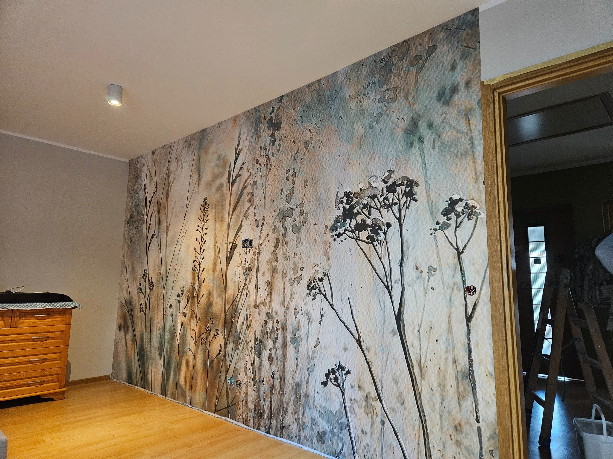 Ściana po położeniu fototapety z motywem polnych traw i kwiatów w odcieniach beżu, brązu i szarości, widoczna drabina malarska i fragment komody z drewna.