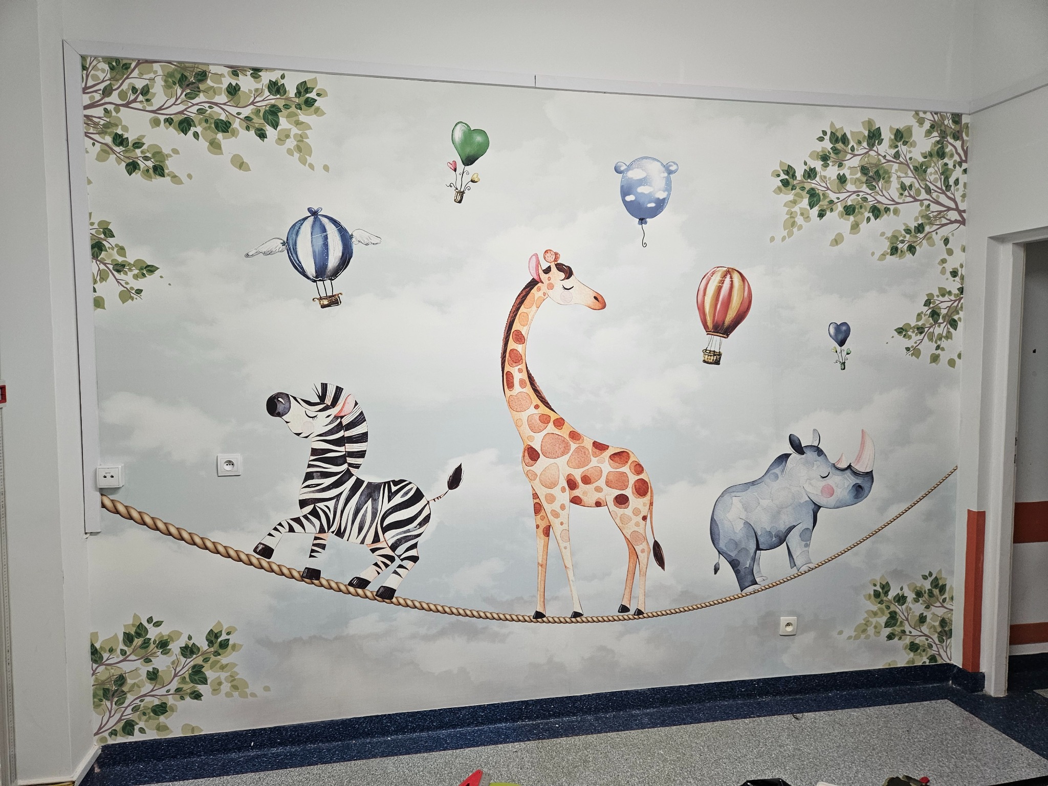 Ściana w pokoju dziecięcym z dekoracyjną tapetą przedstawiającą zebry, żyrafę i nosorożca balansujące na linie, a w tle balony i gałęzie drzew.