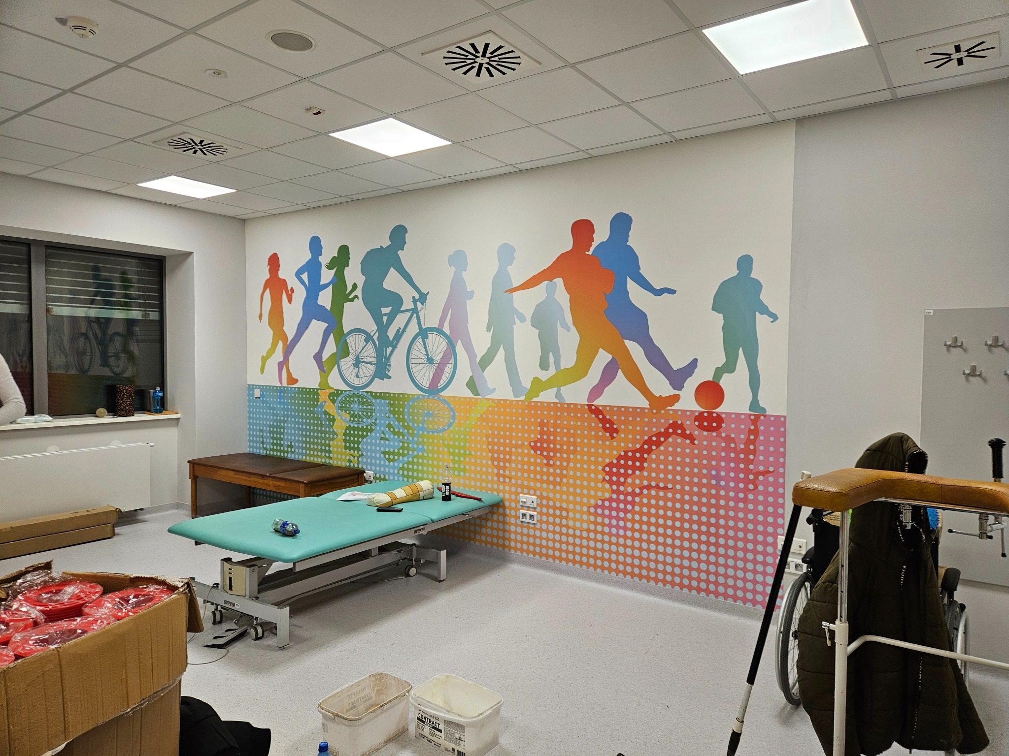 Ściana w gabinecie rehabilitacyjnym ozdobiona barwną grafiką sylwetek sportowców, z widocznym stołem do masażu i sprzętem rehabilitacyjnym.