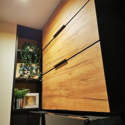 Amadeusz Łacwik Studio Sylvini - Szafa wnękowa z drewnianymi frontami i czarnymi uchwytami, obok półka z dekoracjami i roślinami, widoczny fragment urządzenia AGD z napisem 'ORIGIN INVERTER'.