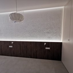 Amadeusz Łacwik Studio Sylvini - Nowoczesna sypialnia z drewnianym panelem ściennym podświetlonym LED, lampa w folii ochronnej, biała ściana i szafa w minimalistycznym stylu.