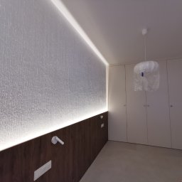 Amadeusz Łacwik Studio Sylvini - Nowoczesna sypialnia z podświetlaną ścianą o teksturze lodu i minimalistyczną zabudową szaf w kolorze białym. Lampa wisząca zabezpieczona folią.