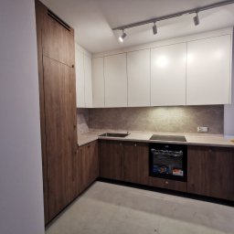 Amadeusz Łacwik Studio Sylvini - Nowoczesna kuchnia z białymi szafkami górnymi i drewnopodobnymi dolnymi, oświetlona reflektorami. Widoczny piekarnik i fragment blatu. Minimalistyczny design.