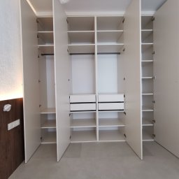 Amadeusz Łacwik Studio Sylvini - Otwarta, biała szafa wnękowa z półkami i szufladami, widok z przodu. Minimalistyczny design, jasne wnętrze, drewniane wykończenie ściany po lewej stronie.