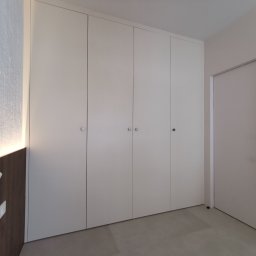 Amadeusz Łacwik Studio Sylvini - Biała, czterodrzwiowa szafa wnękowa z okrągłymi uchwytami, w minimalistycznym wnętrzu z szarą podłogą i oświetleniem LED na brązowej ściance.