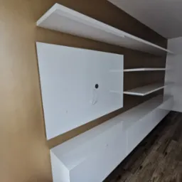 Biała, nowoczesna meblościanka z półkami i szafkami RTV na tle brązowej ściany. Widoczne drewniane panele podłogowe i fragment okna.