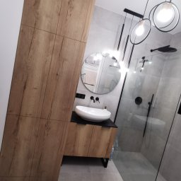 Amadeusz Łacwik Studio Sylvini - Nowoczesna łazienka z drewnianą szafką pod umywalką nablatową, szarą glazurą i kabiną prysznicową typu walk-in. Okrągłe lustro i designerskie oświetlenie.