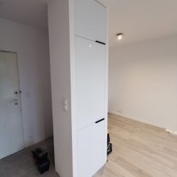 Amadeusz Łacwik Studio Sylvini - Biała szafa wnękowa z czarnymi uchwytami, wkomponowana w ścianę, obok widoczna skrzynka narzędziowa. Jasne panele na podłodze.