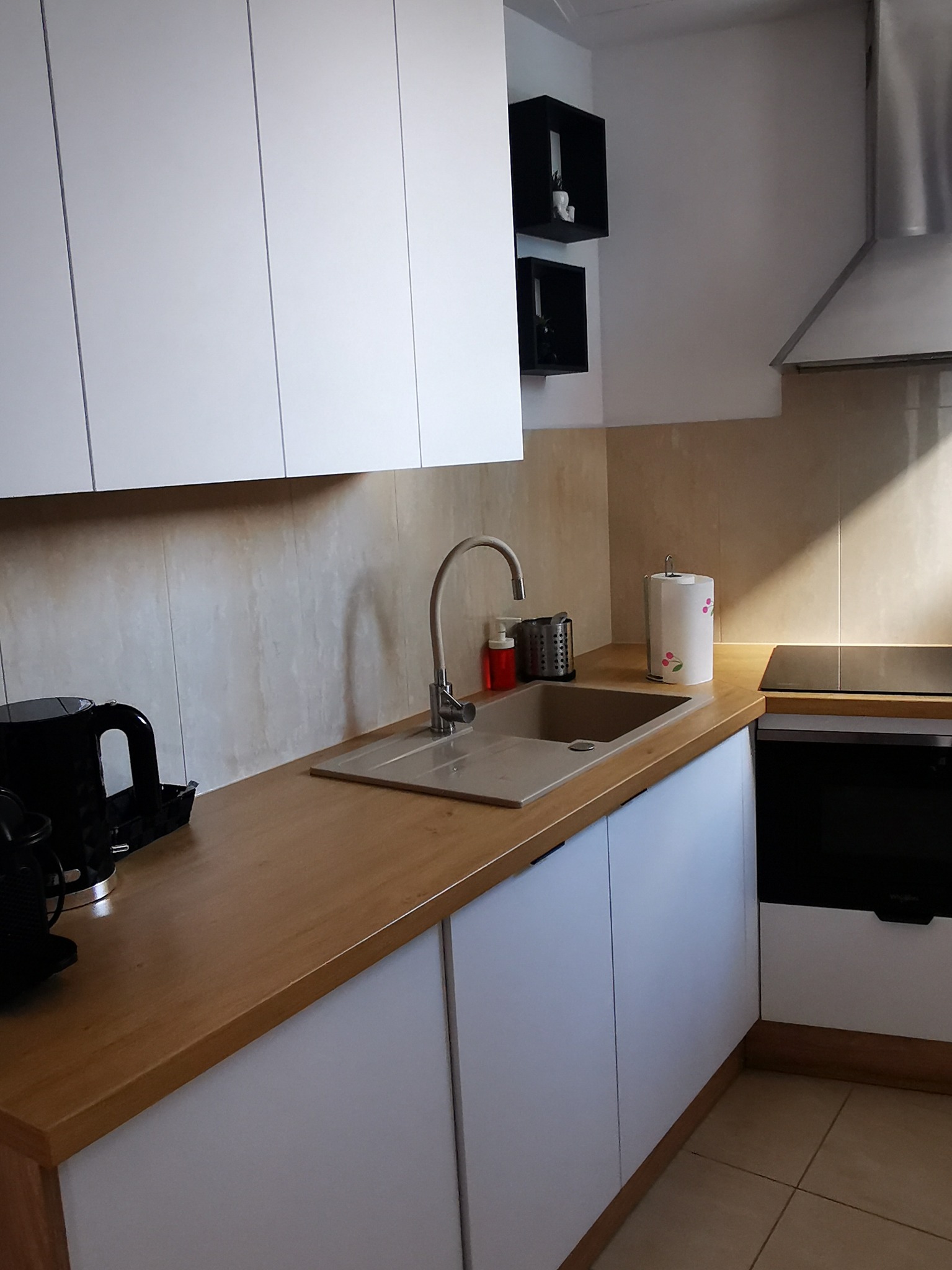 Nowoczesna kuchnia z białymi szafkami, drewnianym blatem, szarym zlewozmywakiem i minimalistycznymi czarnymi półkami na ścianie.