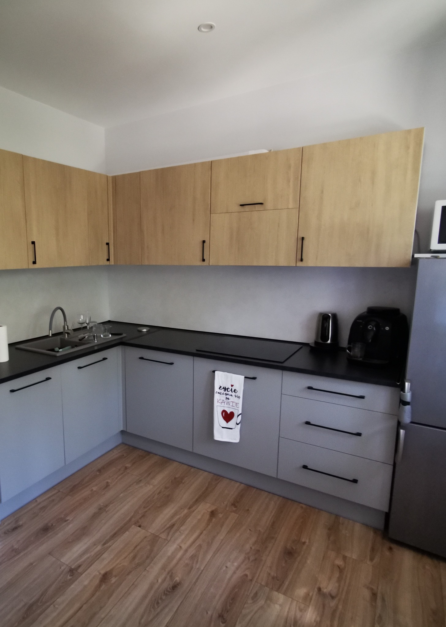 Jasna kuchnia z szarymi szafkami dolnymi i drewnopodobnymi górnymi, czarnym blatem, widoczne akcesoria kuchenne i ręcznik z motywem kawy.