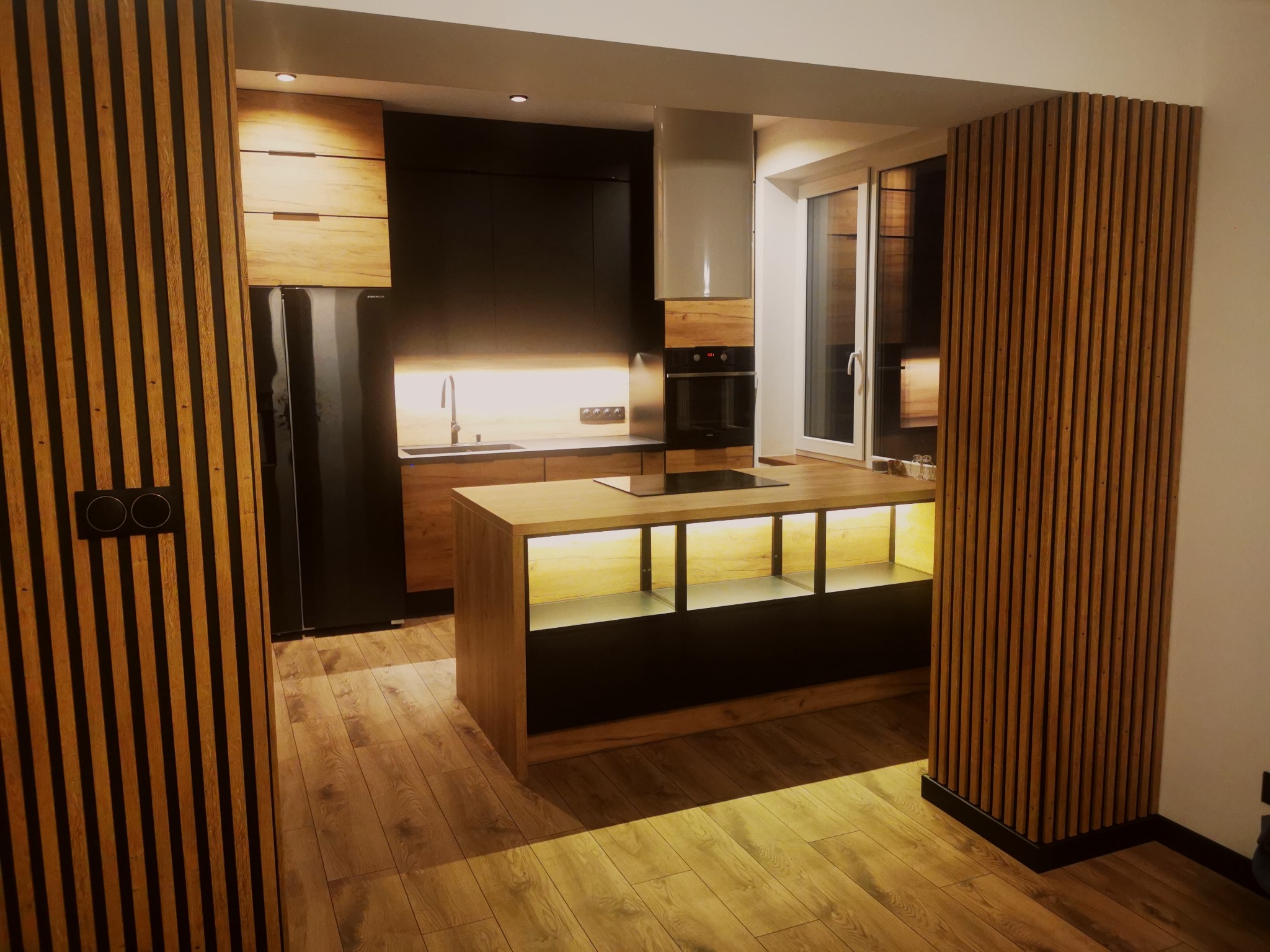 Nowoczesna kuchnia z wyspą, fronty w odcieniach drewna i czerni, oświetlenie LED pod wyspą, drewniane lamele dekoracyjne przy wejściu i oknie.
