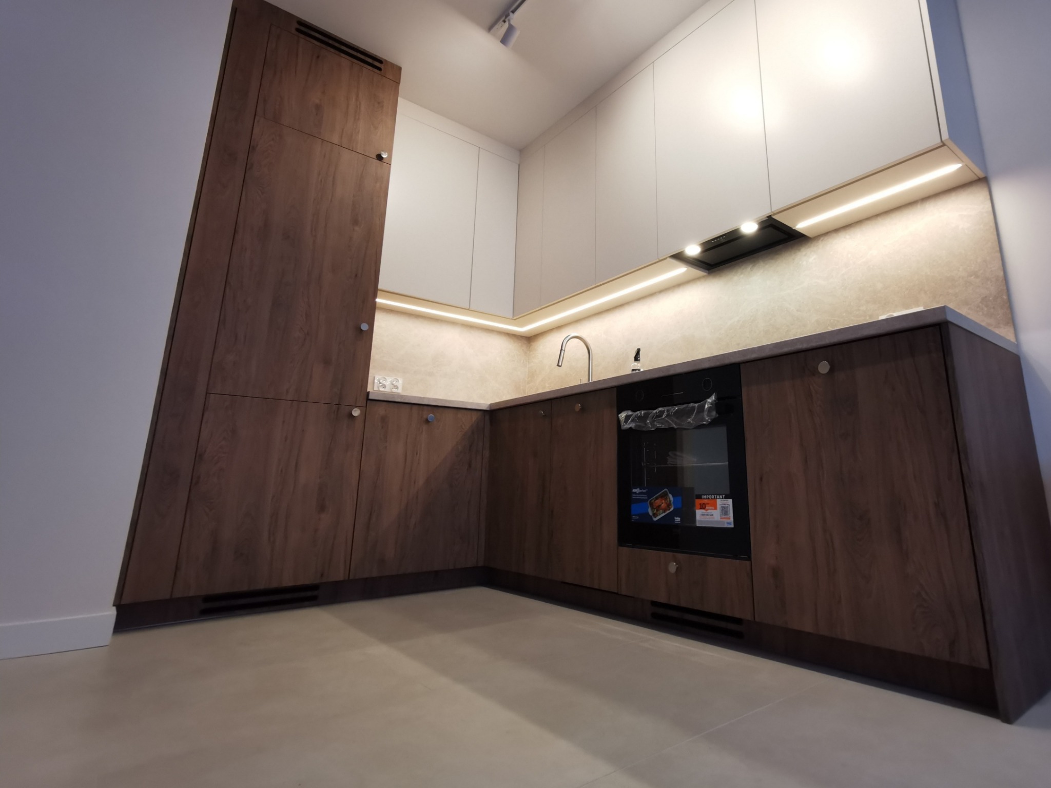 Nowoczesna kuchnia na wymiar z drewnopodobnymi frontami i białymi szafkami wiszącymi, oświetlona ledami pod szafkami. Widok z niskiego kąta.