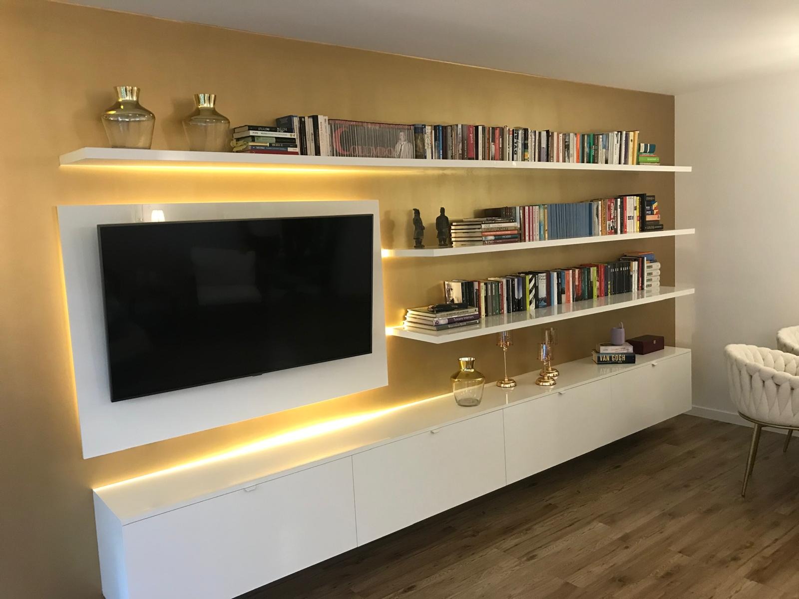 Nowoczesna zabudowa ściany z białymi szafkami, półkami i podświetleniem LED. Na półkach książki i dekoracje na tle złotej ściany.