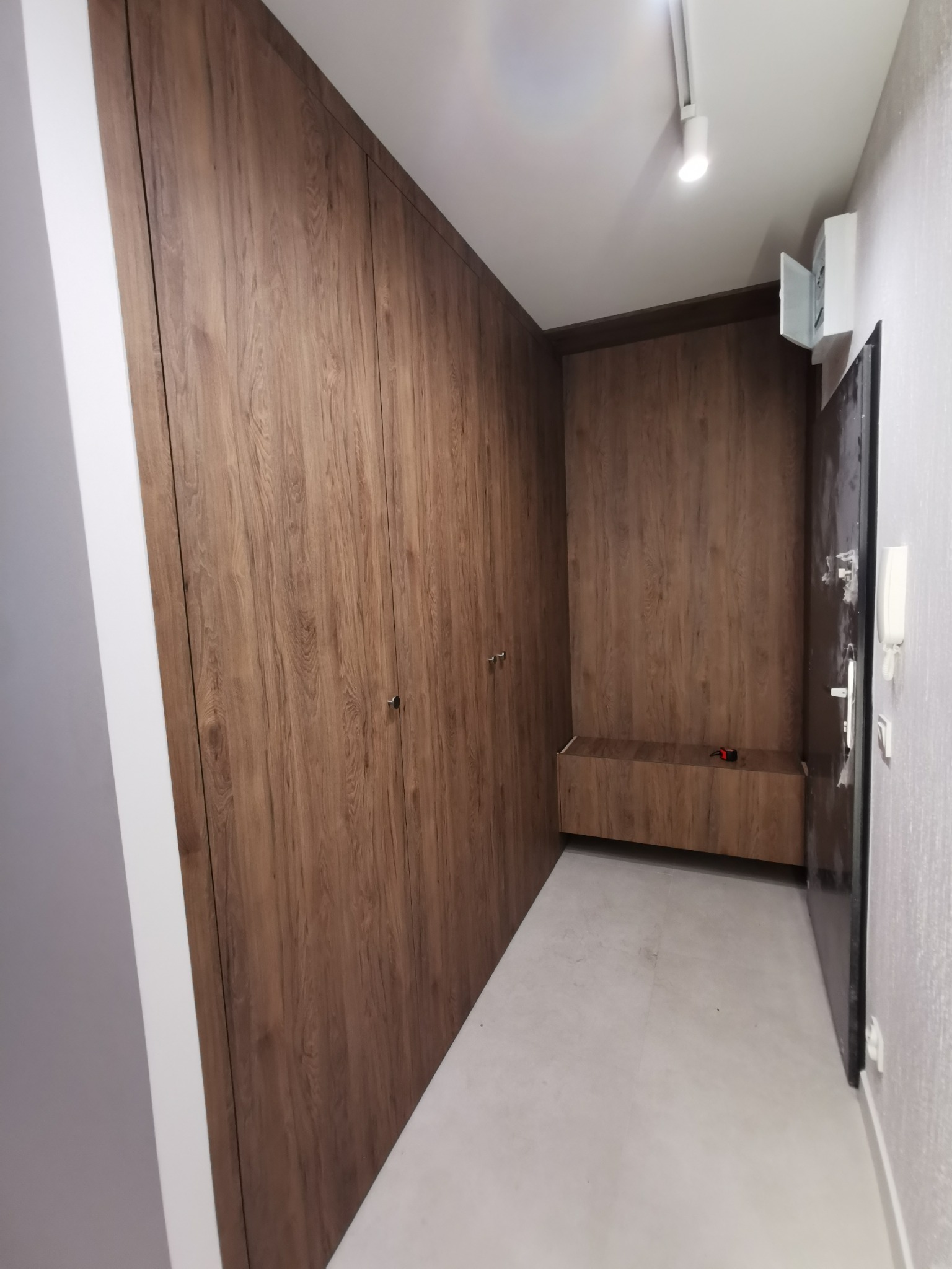 Wnętrze przedpokoju z zabudowaną szafą w odcieniach drewna, z wbudowanym siedziskiem. Minimalistyczny design, oświetlenie punktowe, widoczny fragment drzwi wejściowych.