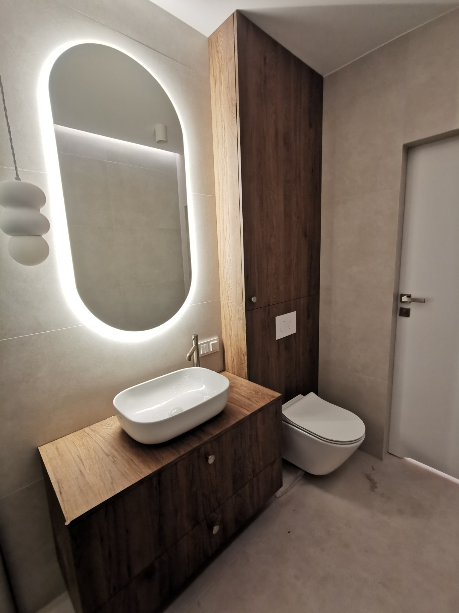 Nowoczesna łazienka z drewnianą szafką pod umywalką, owalnym lustrem z podświetleniem LED i zabudową meblową z ciemnego drewna.
