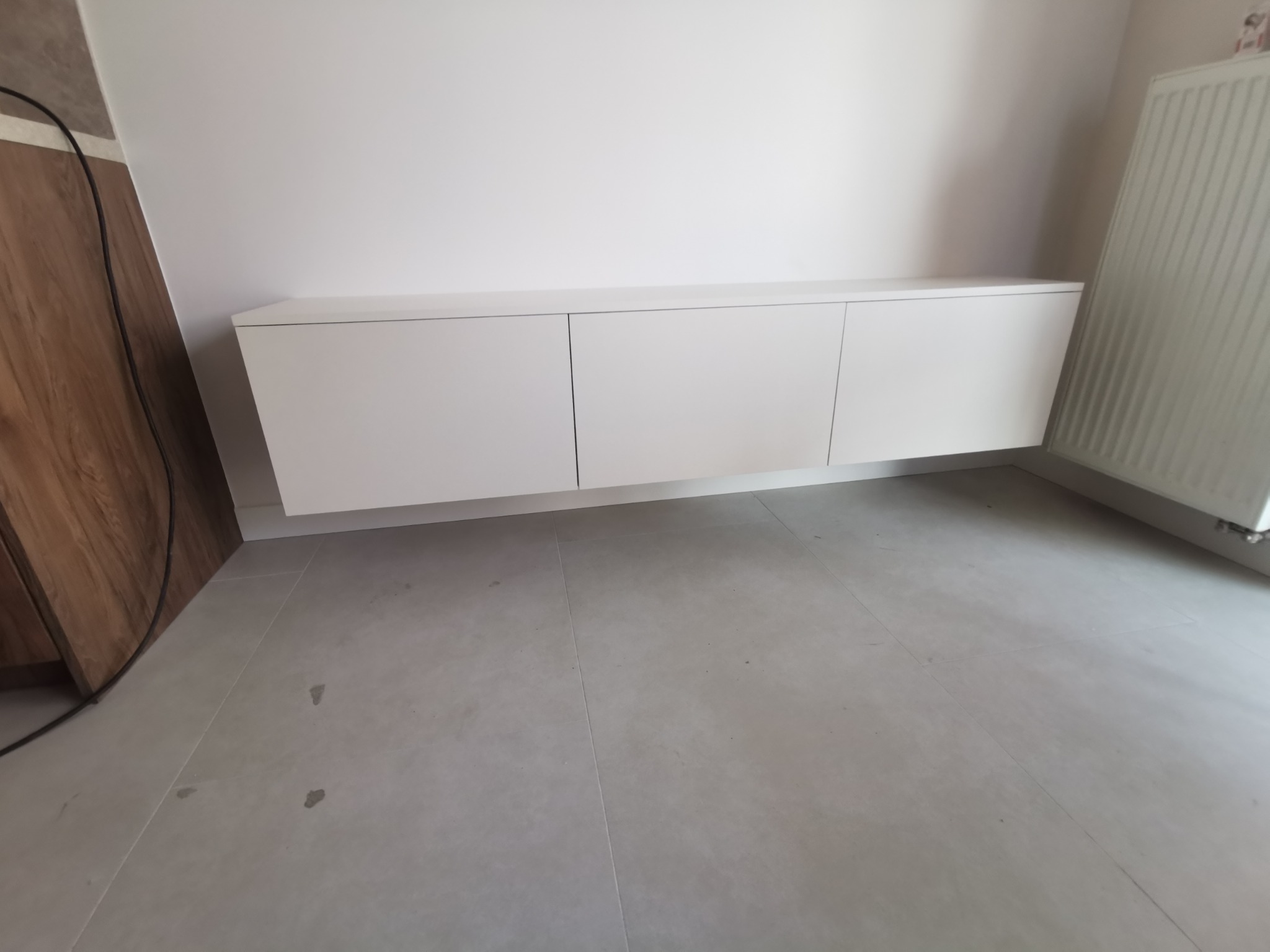 Biała, podwieszana szafka RTV z trzema frontami, zamontowana na ścianie w nowoczesnym wnętrzu. Widoczny fragment drewnianej zabudowy i grzejnik.