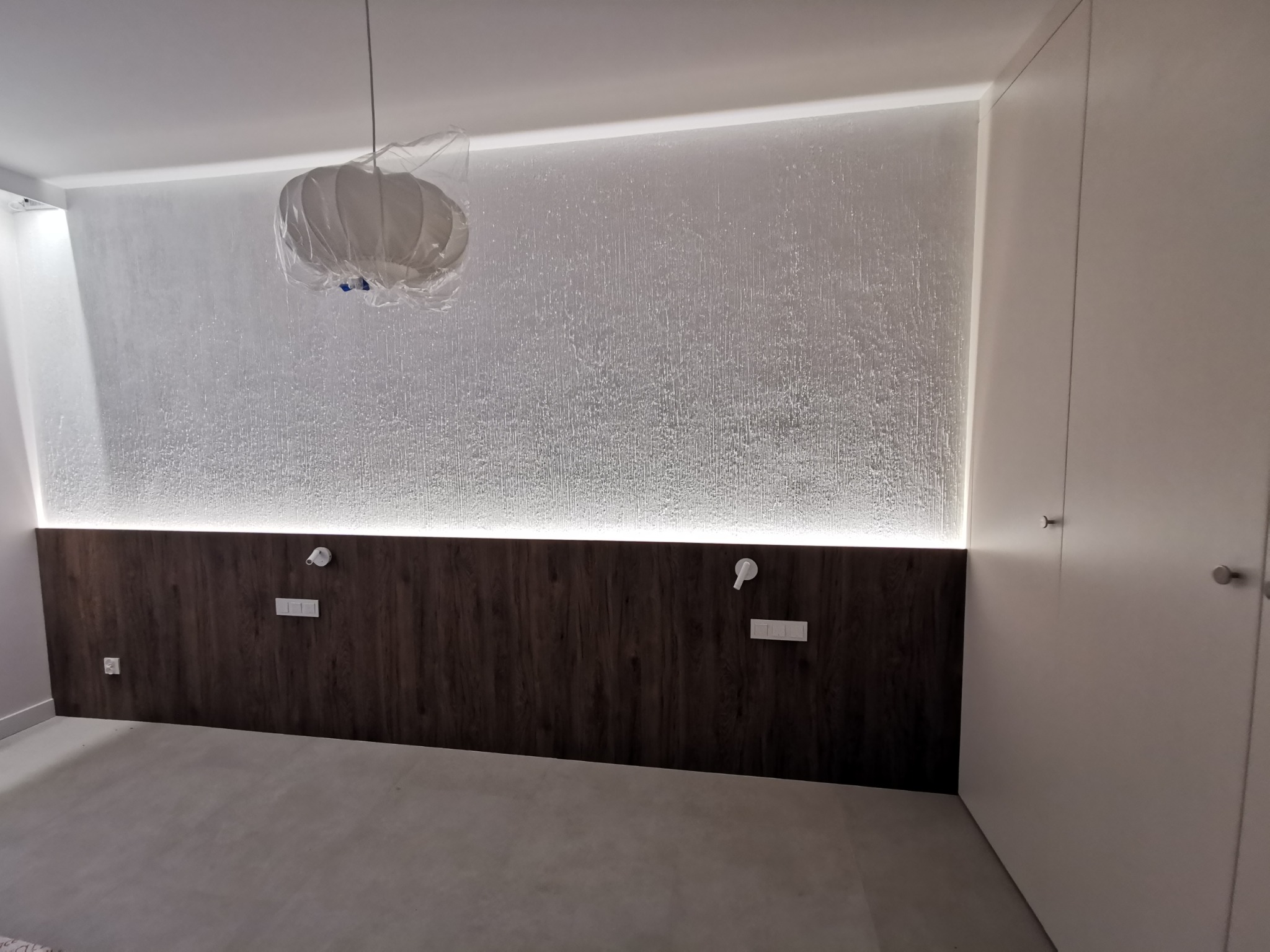 Nowoczesna sypialnia z drewnianym panelem ściennym podświetlonym LED, lampa w folii ochronnej, biała ściana i szafa w minimalistycznym stylu.