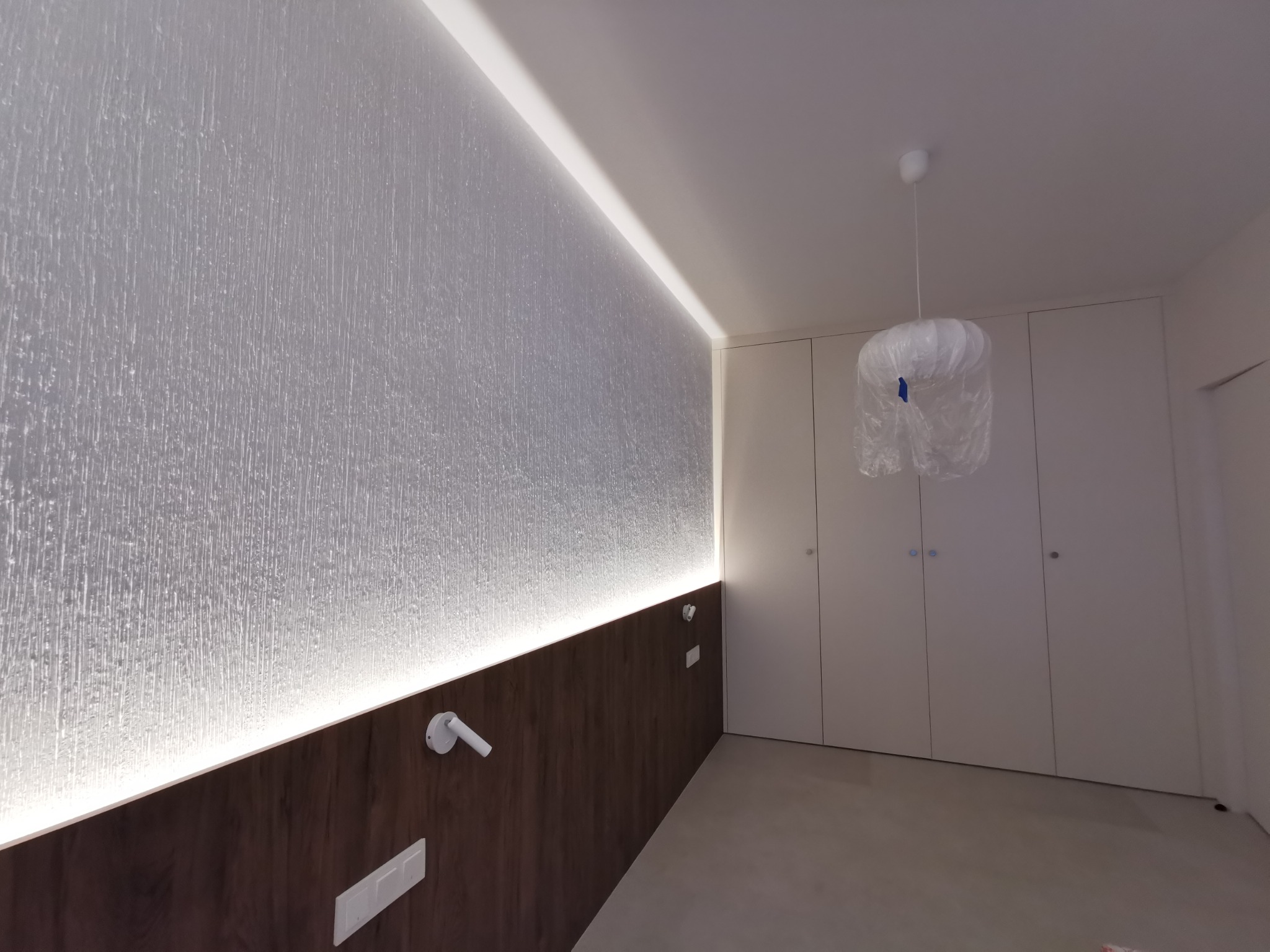 Nowoczesna sypialnia z podświetlaną ścianą o teksturze lodu i minimalistyczną zabudową szaf w kolorze białym. Lampa wisząca zabezpieczona folią.