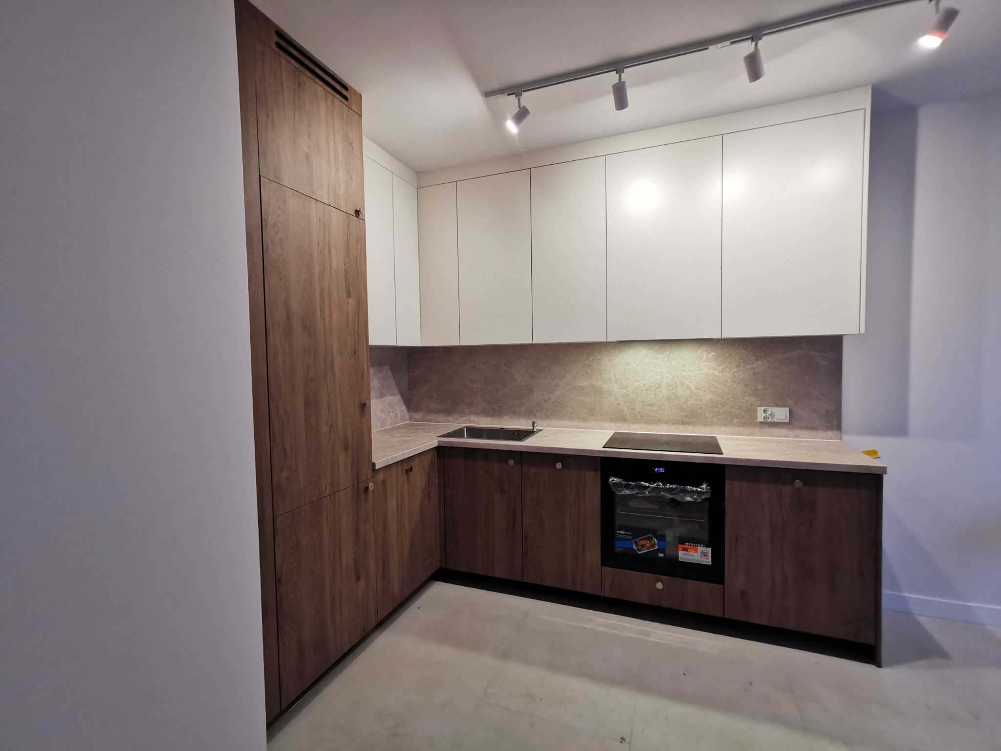 Nowoczesna kuchnia z białymi szafkami górnymi i drewnopodobnymi dolnymi, oświetlona reflektorami. Widoczny piekarnik i fragment blatu. Minimalistyczny design.