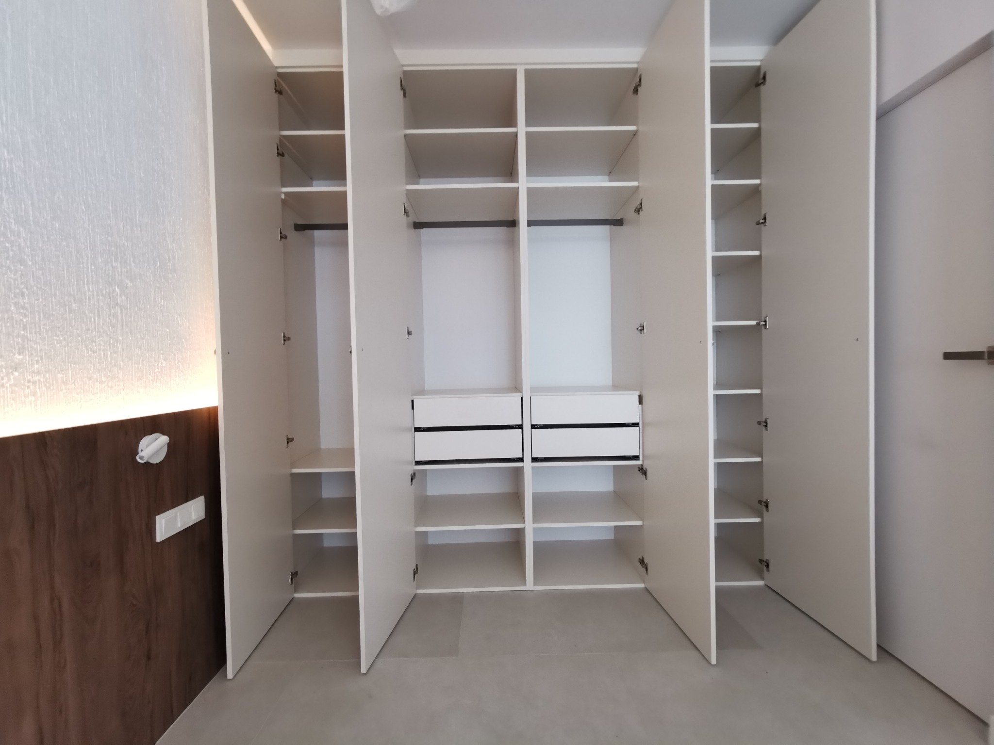 Otwarta, biała szafa wnękowa z półkami i szufladami, widok z przodu. Minimalistyczny design, jasne wnętrze, drewniane wykończenie ściany po lewej stronie.