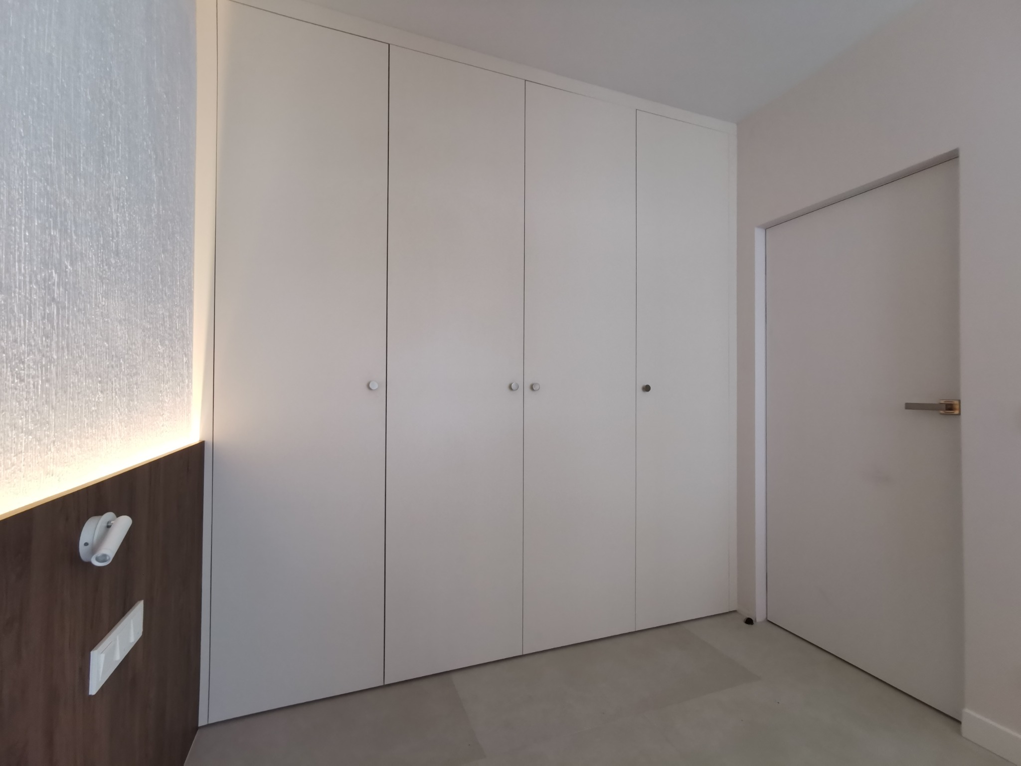 Biała, czterodrzwiowa szafa wnękowa z okrągłymi uchwytami, w minimalistycznym wnętrzu z szarą podłogą i oświetleniem LED na brązowej ściance.