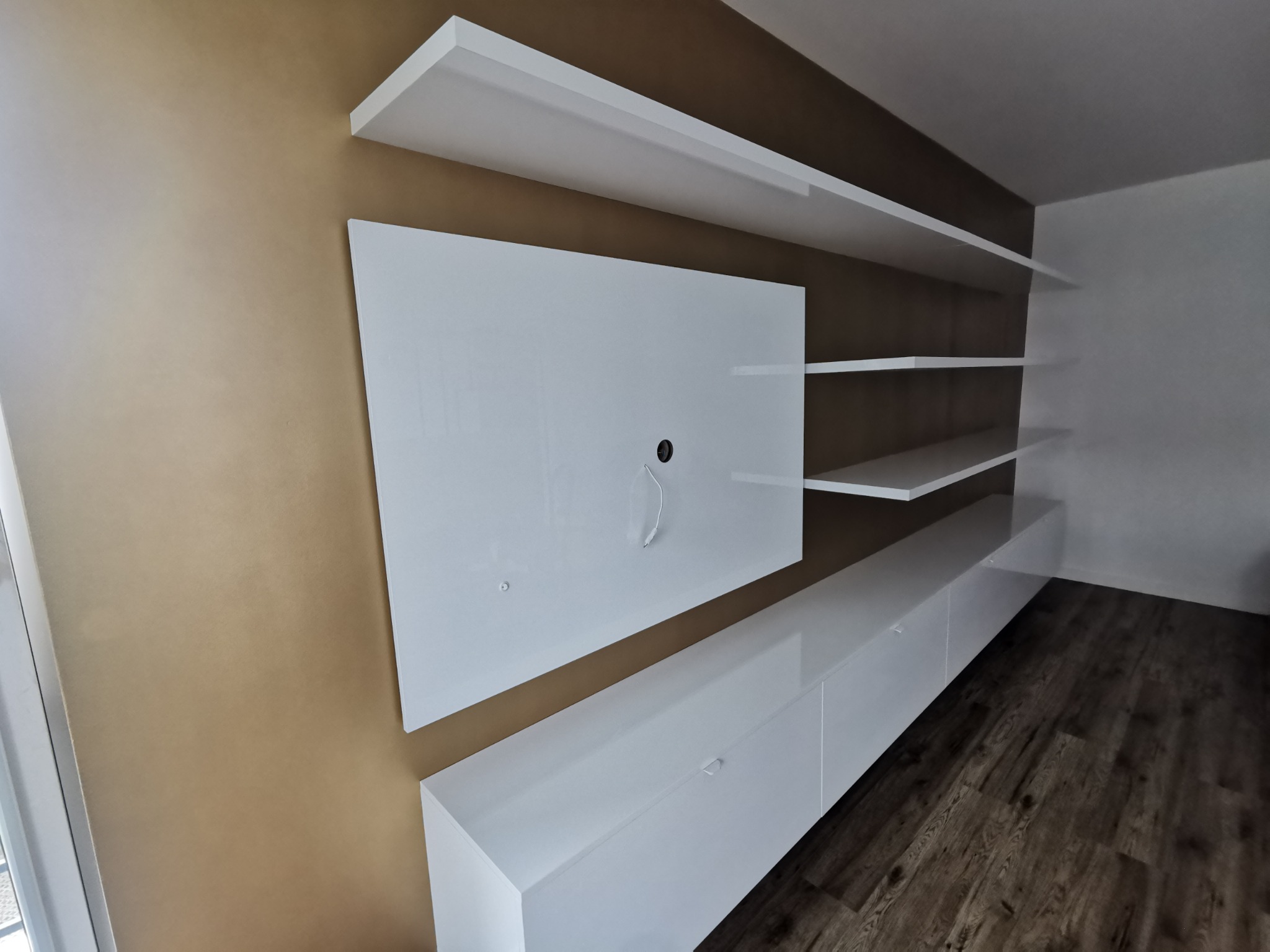 Biała, nowoczesna meblościanka z półkami i szafkami RTV na tle brązowej ściany. Widoczne drewniane panele podłogowe i fragment okna.