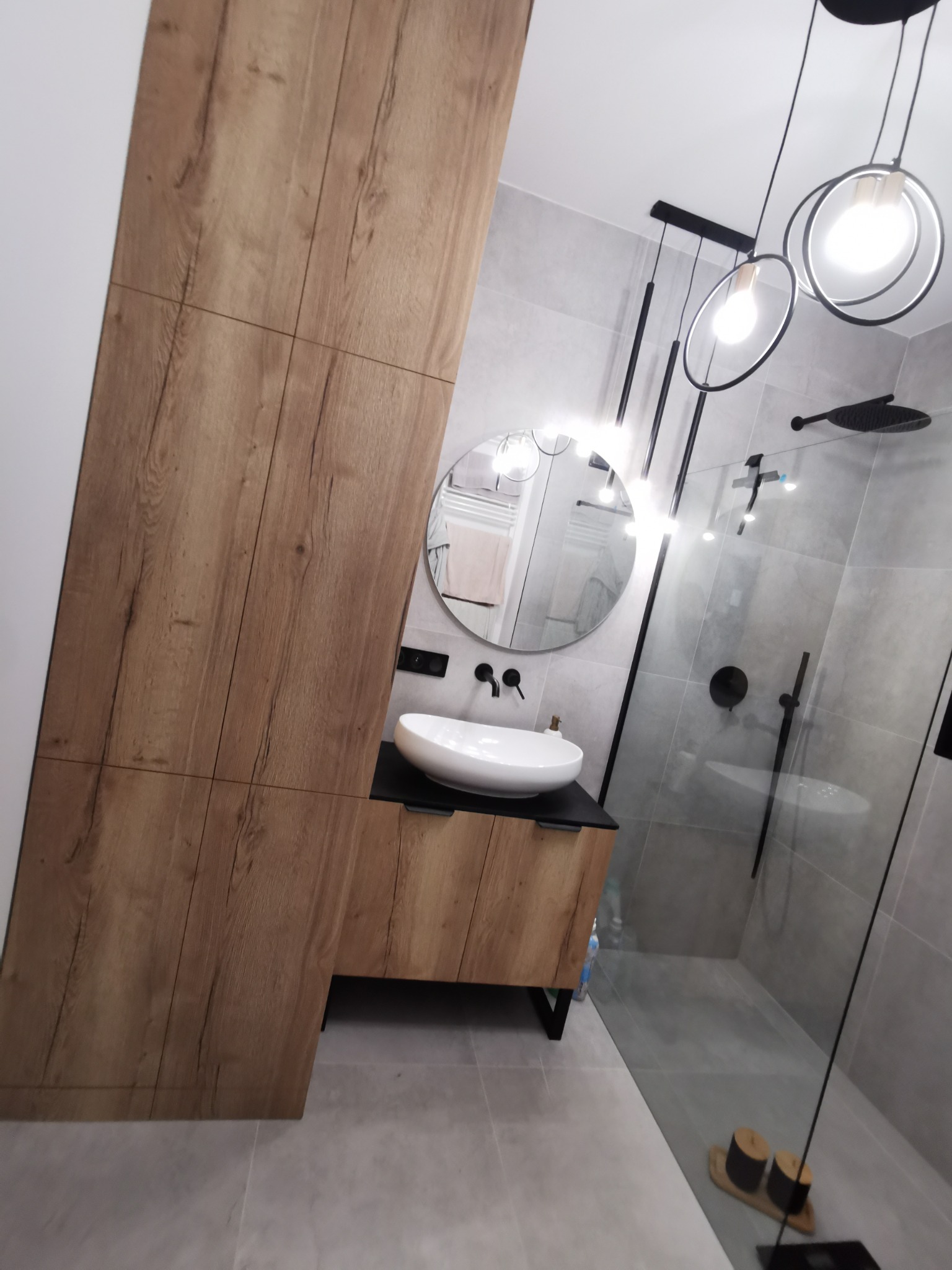 Nowoczesna łazienka z drewnianą szafką pod umywalką nablatową, szarą glazurą i kabiną prysznicową typu walk-in. Okrągłe lustro i designerskie oświetlenie.