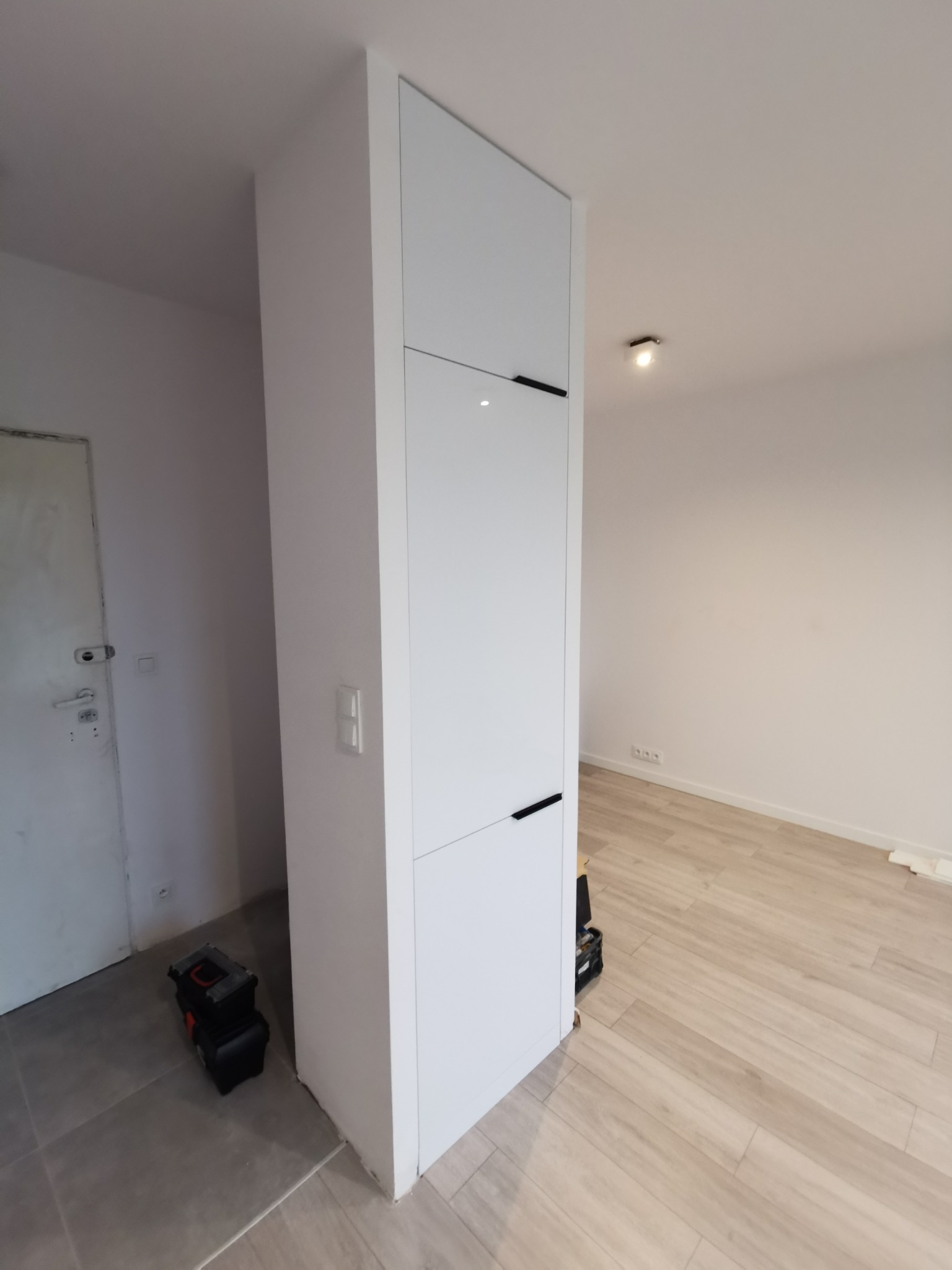 Biała szafa wnękowa z czarnymi uchwytami, wkomponowana w ścianę, obok widoczna skrzynka narzędziowa. Jasne panele na podłodze.