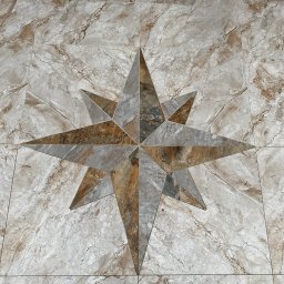 Marek Jarosz ZAKŁAD REMONTOWO-BUDOWLANY - Wzorzysta podłoga z płytek imitujących marmur, z centralnie umieszczoną dekoracją w kształcie gwiazdy, wykonaną z płytek w odcieniach szarości i brązu. Widok z góry.