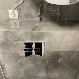 Marek Jarosz ZAKŁAD REMONTOWO-BUDOWLANY - Szara, błyszcząca ściana w łazience z chromowanym przyciskiem spłuczki Roca, widoczne zawory i gniazdko elektryczne. Minimalistyczny design.