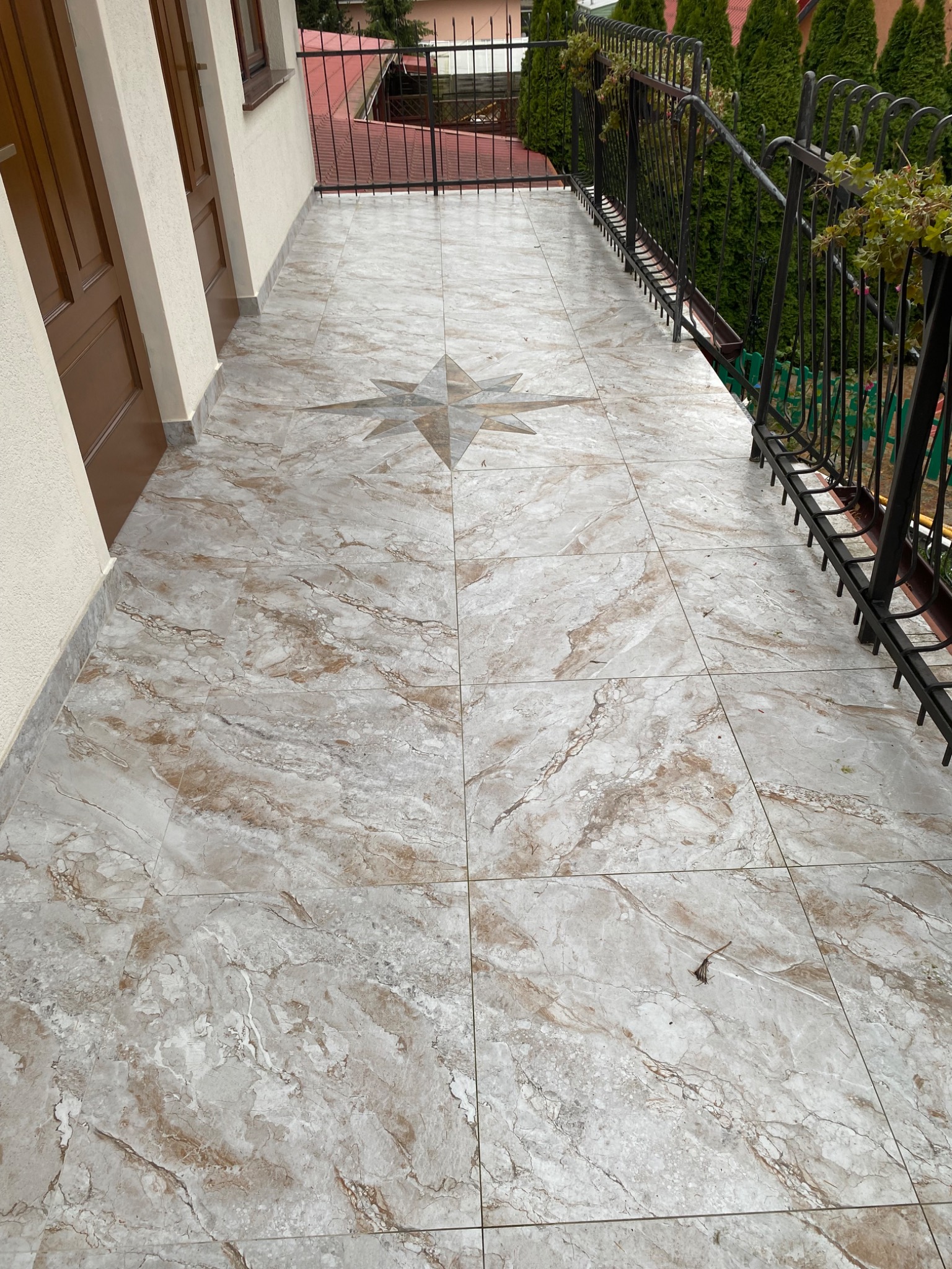 Wykończony balkon z marmurową posadzką w odcieniach beżu i szarości, wzór gwiazdy na środku. Metalowa balustrada z zielenią w tle. Widok z góry, naturalne światło.