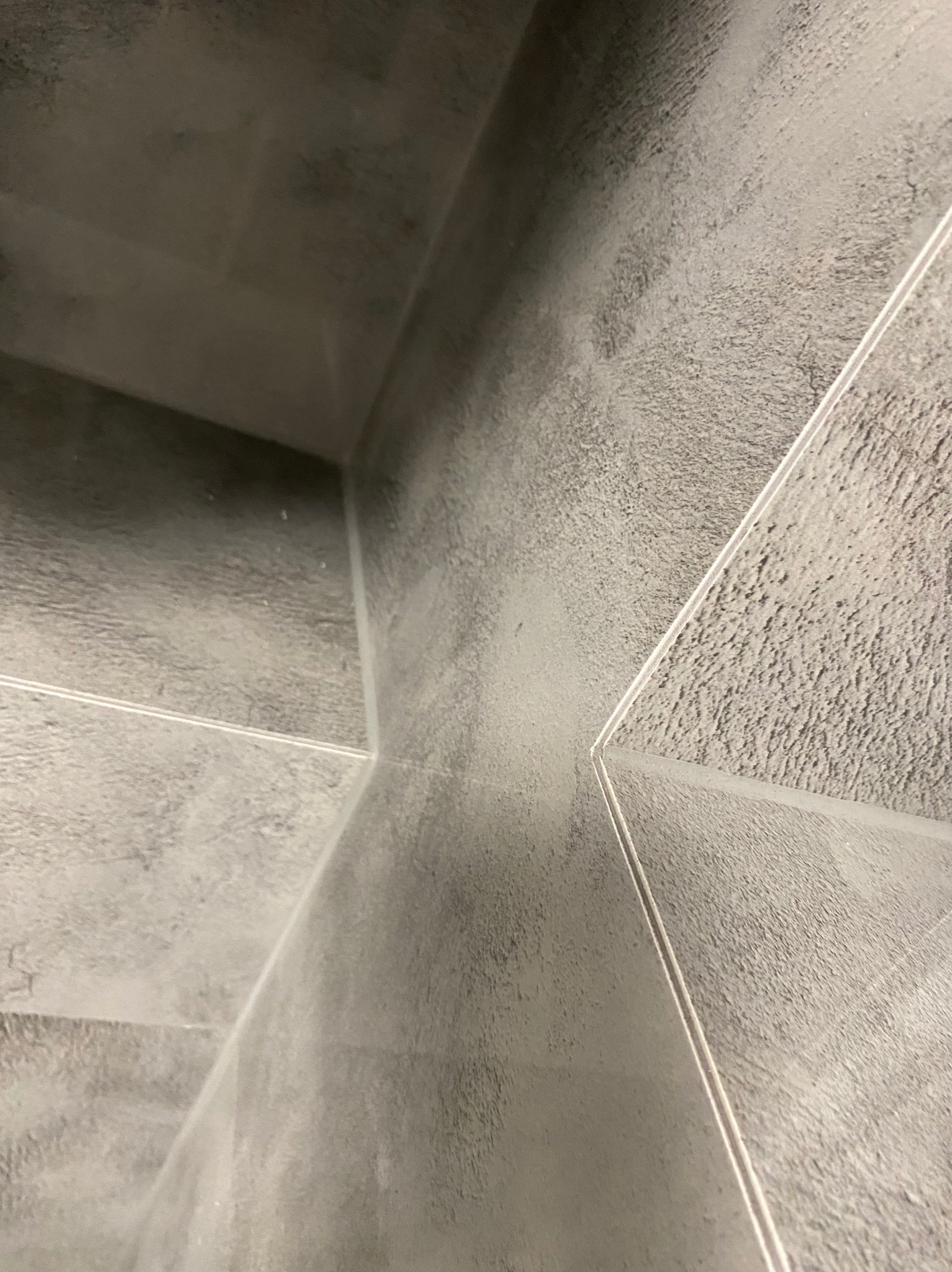 Szare, matowe płytki ceramiczne ułożone pod kątem, z widocznymi fugami. Minimalistyczny design, surowa estetyka, bliski kadr na fragment ściany lub podłogi.