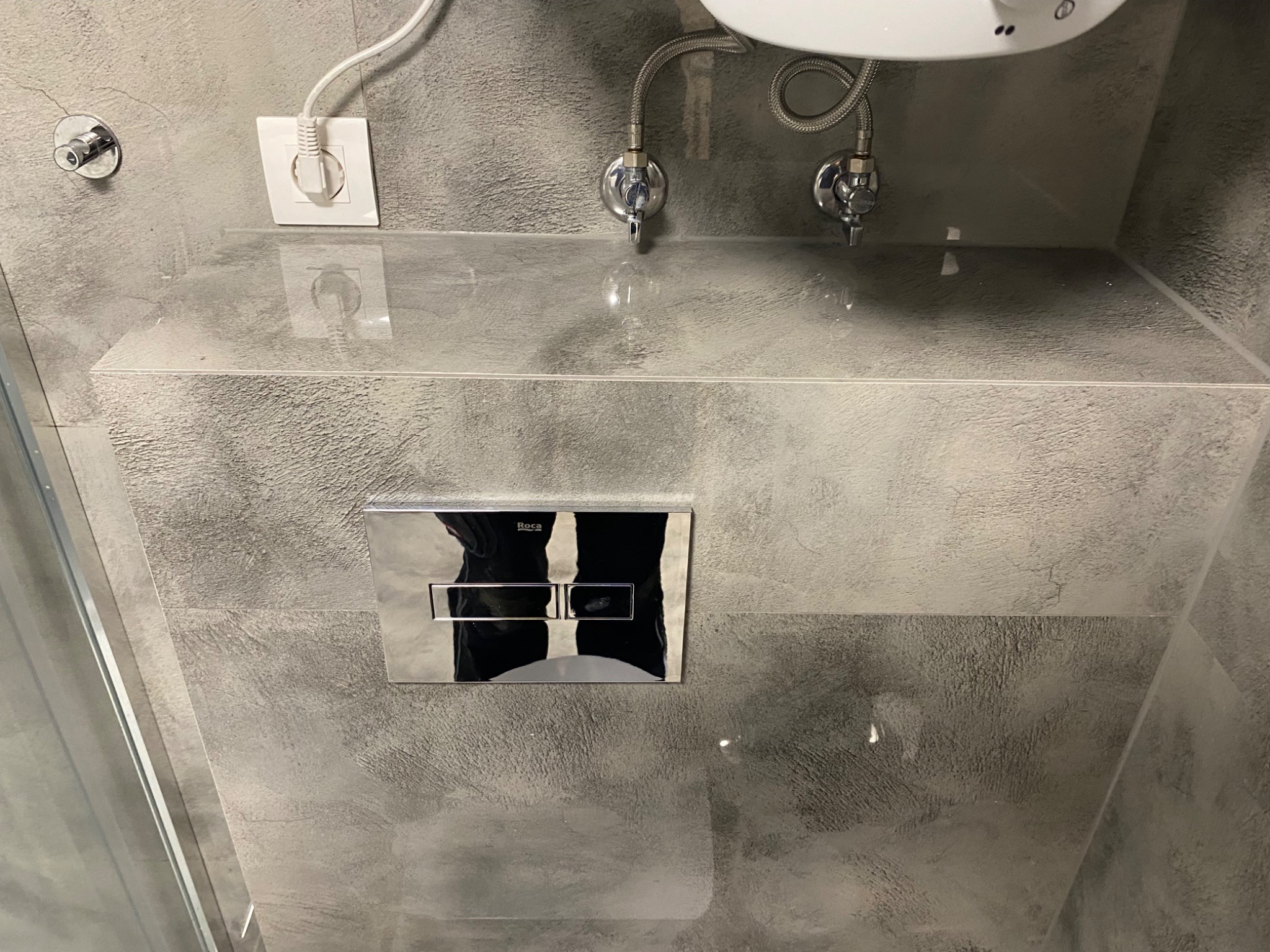 Szara, błyszcząca ściana w łazience z chromowanym przyciskiem spłuczki Roca, widoczne zawory i gniazdko elektryczne. Minimalistyczny design.