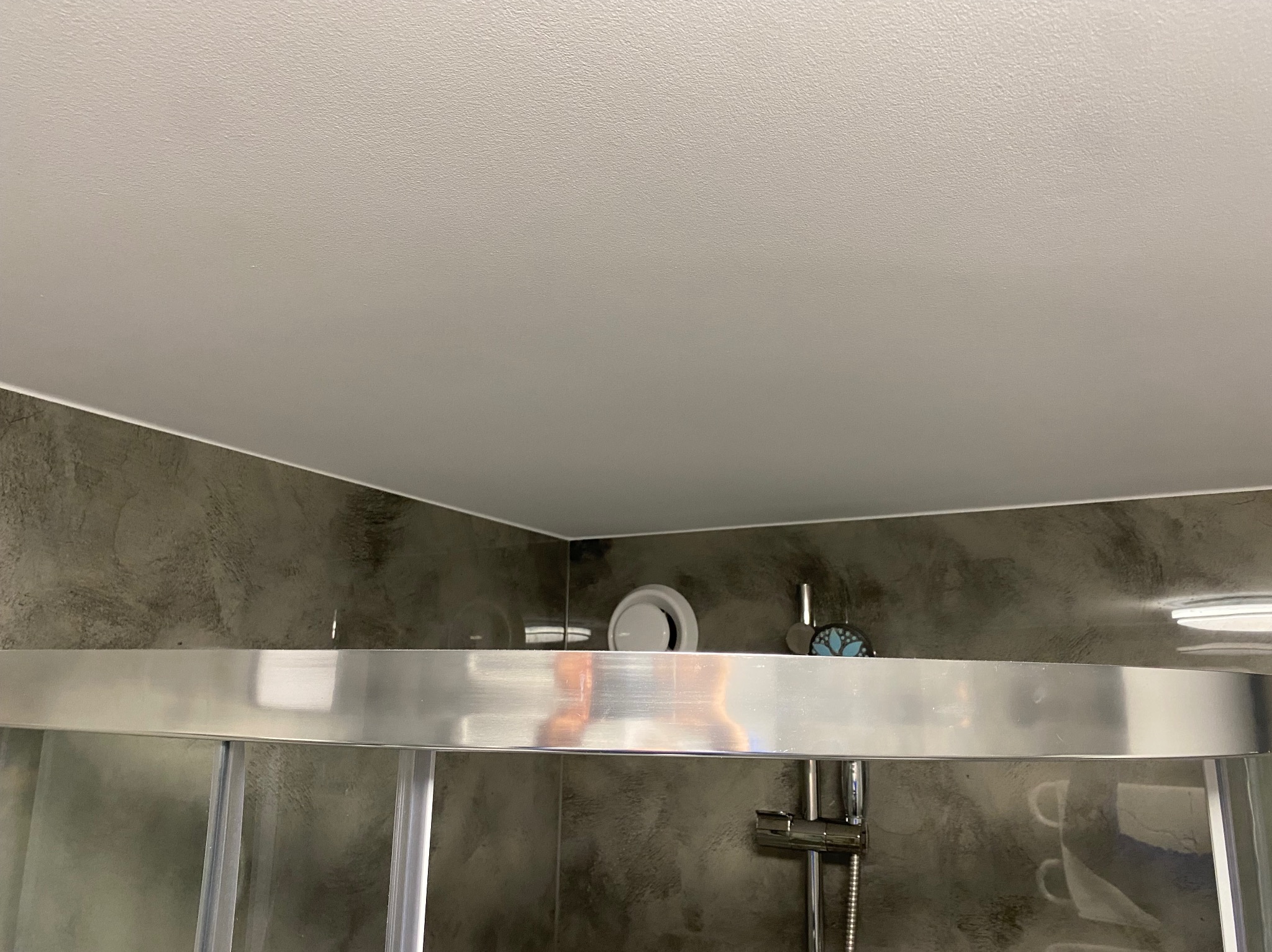 Szczegółowe ujęcie kabiny prysznicowej z szarą, teksturowaną ścianą i białym sufitem. Widoczny wentylator sufitowy i metalowe elementy kabiny.