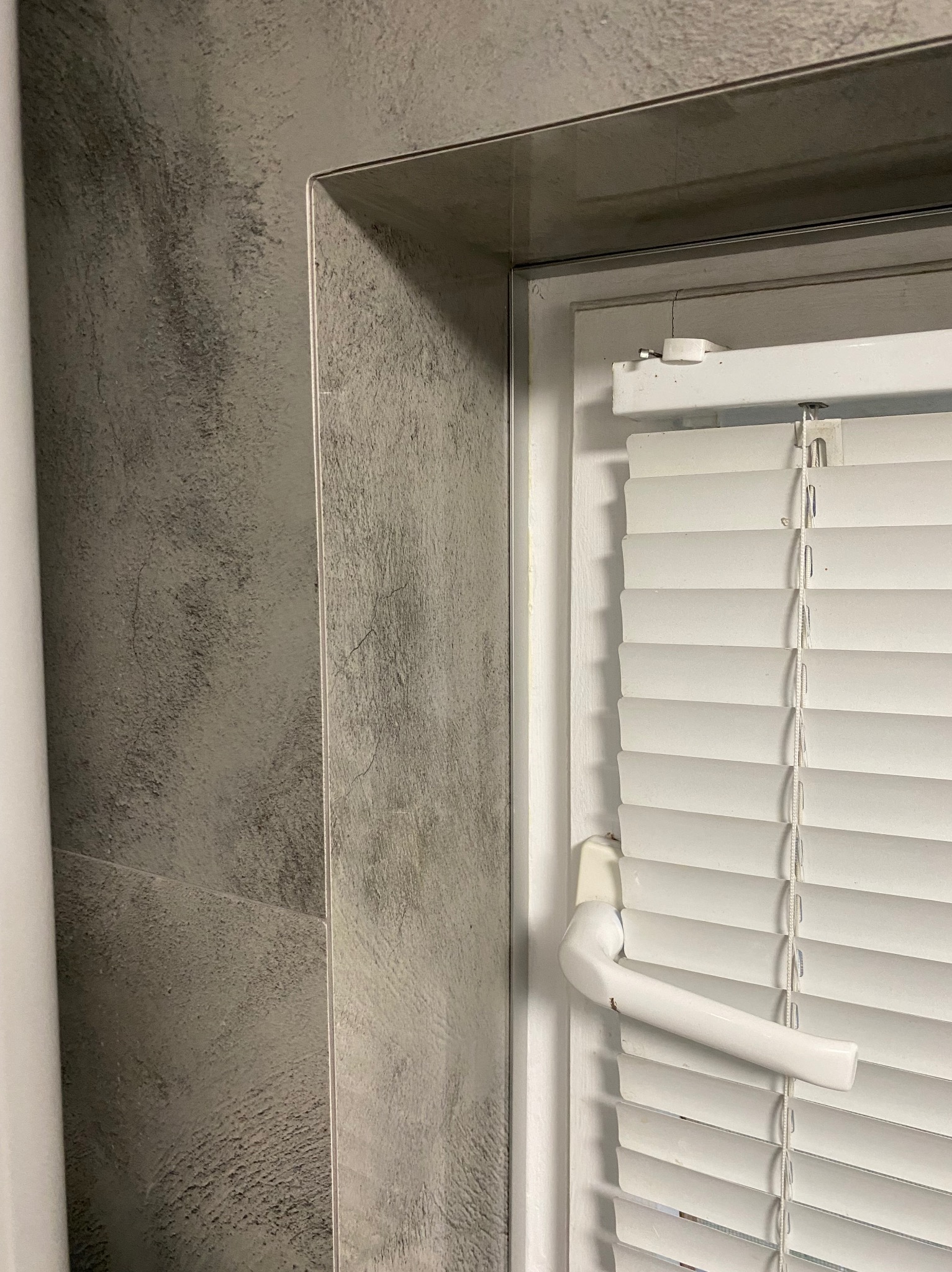 Szare płytki imitujące beton i białe żaluzje okienne w łazience po remoncie w Gryficach. Widoczny fragment ramy okiennej i uchwyt żaluzji.