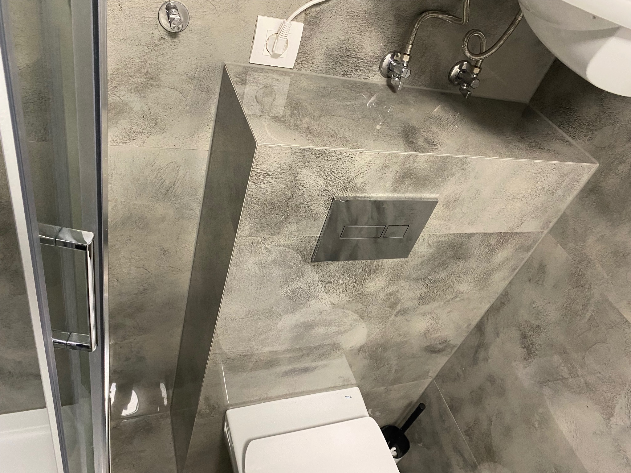 Szara łazienka z toaletą podwieszaną i kabiną prysznicową. Ściany wykończone płytkami imitującymi beton. Widoczny fragment instalacji wodnej i gniazdko elektryczne.