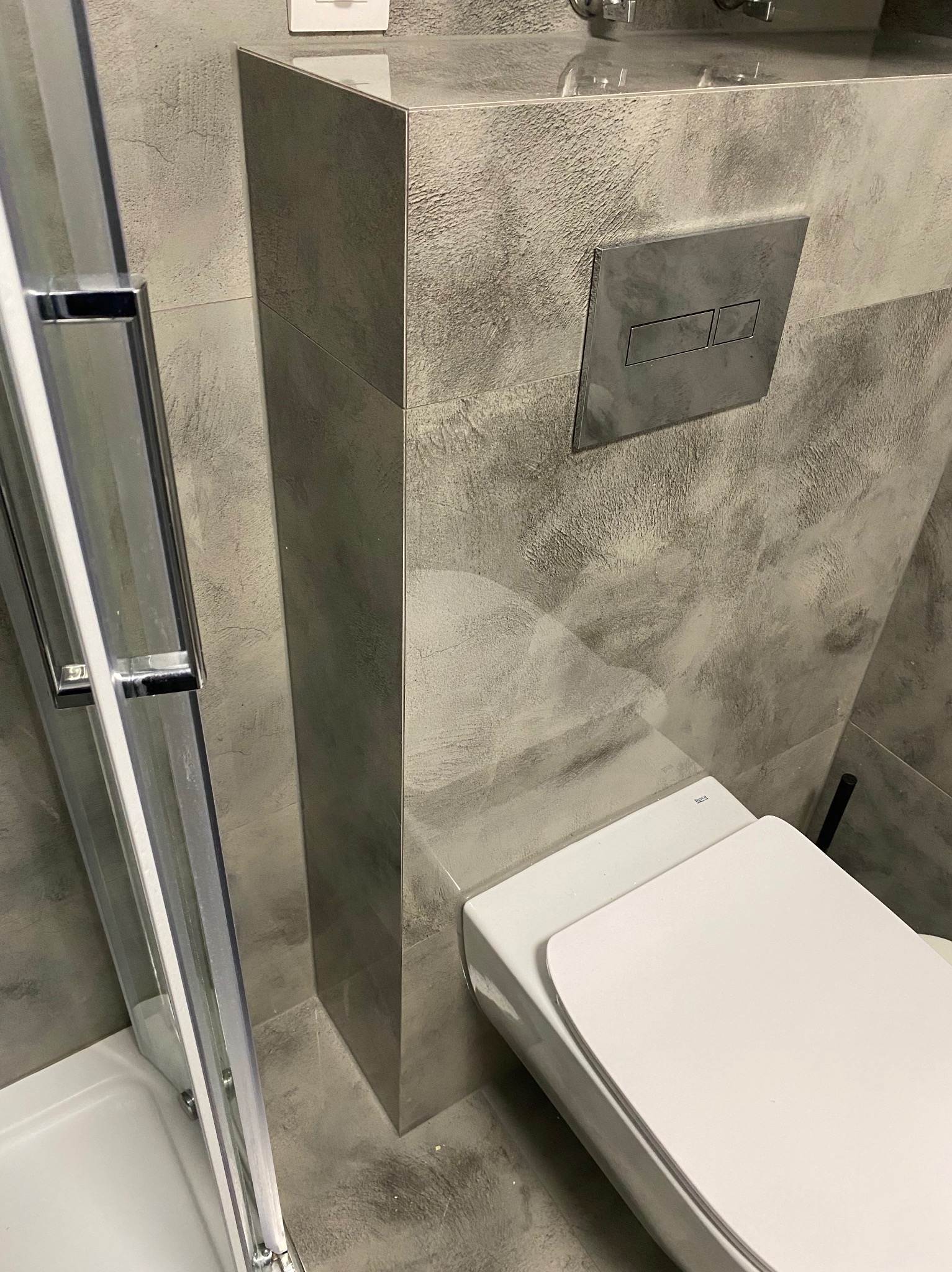 Szara łazienka z podwieszaną toaletą i kabiną prysznicową. Ściany wykończone płytkami imitującymi beton. Minimalistyczny design, nowoczesne wykończenie.