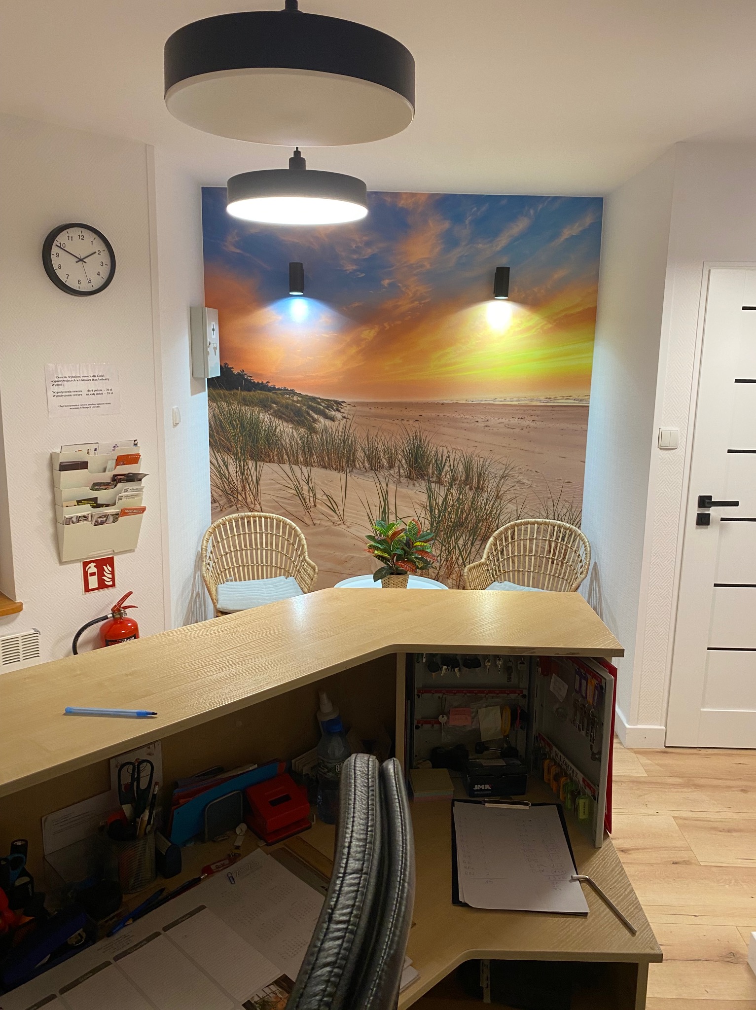 Recepcja z fototapetą plaży o zachodzie słońca, rattanowe krzesła, drewniana podłoga i designerskie lampy sufitowe. Przytulne wnętrze biura.