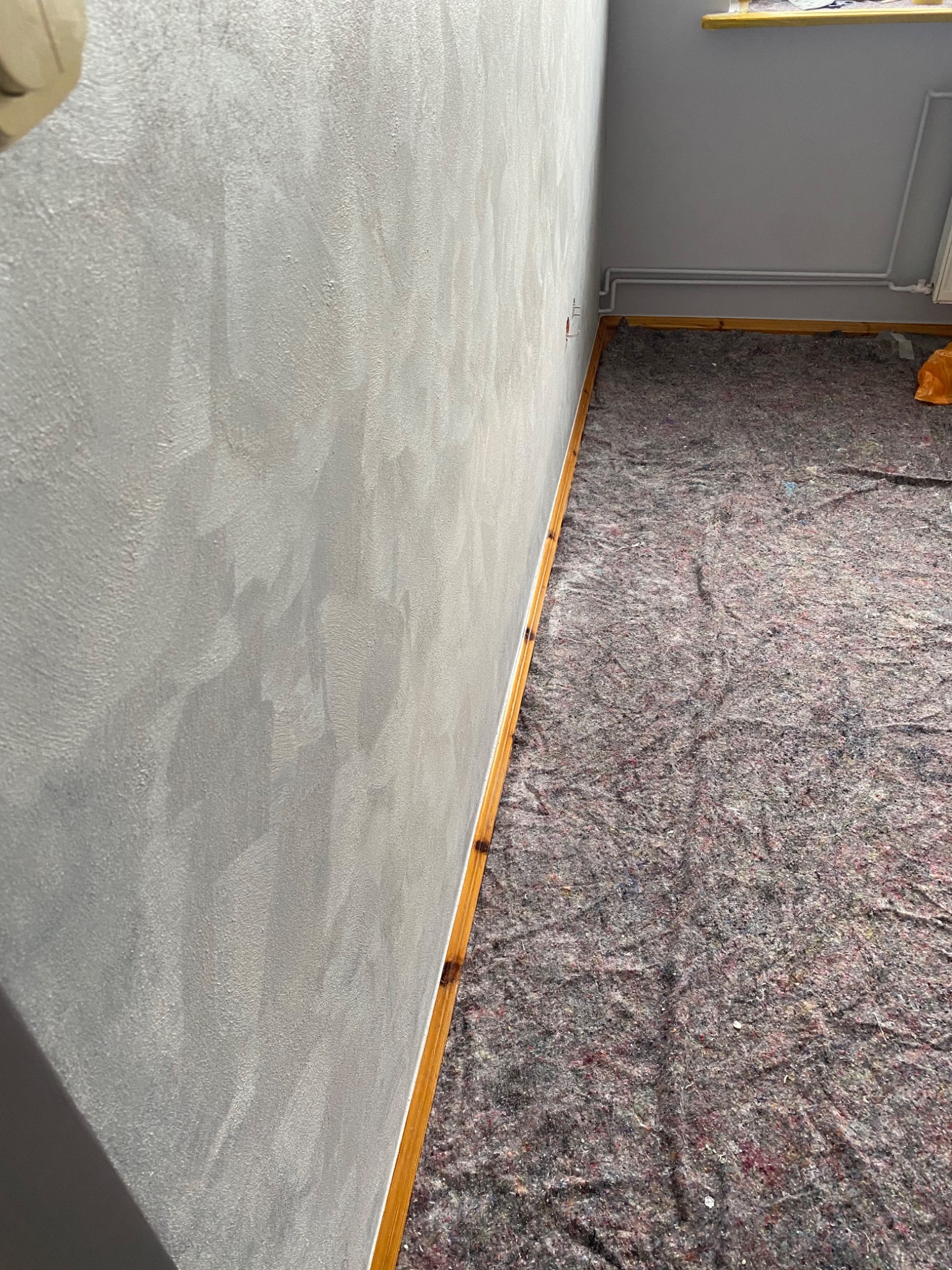 Ściana po malowaniu dekoracyjnym w odcieniach szarości, zabezpieczona podłoga z listwami przypodłogowymi. Widok z dołu, ukazujący fakturę ściany i przygotowanie do dalszych prac.