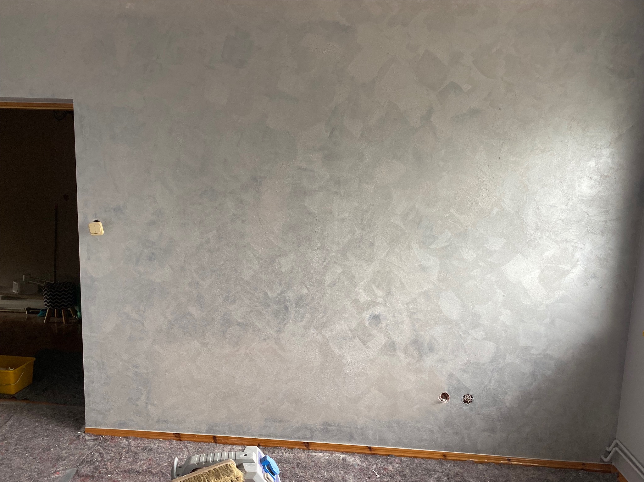 Ściana z efektem dekoracyjnym w odcieniach szarości i srebra, widoczne gniazdka elektryczne i fragment podłogi zabezpieczonej folią malarską. Widok na otwarte drzwi.