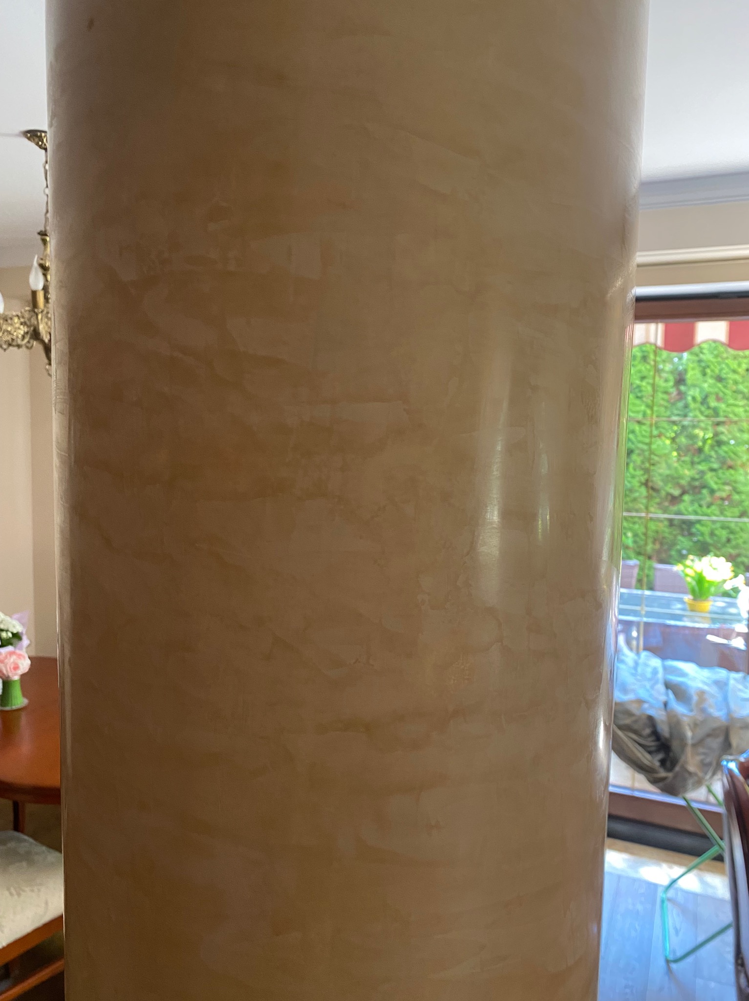 Dekoracyjna kolumna wewnątrz pomieszczenia, wykończona beżowym tynkiem strukturalnym z efektem marmuru. W tle widoczne okno i fragment wyposażenia wnętrza.