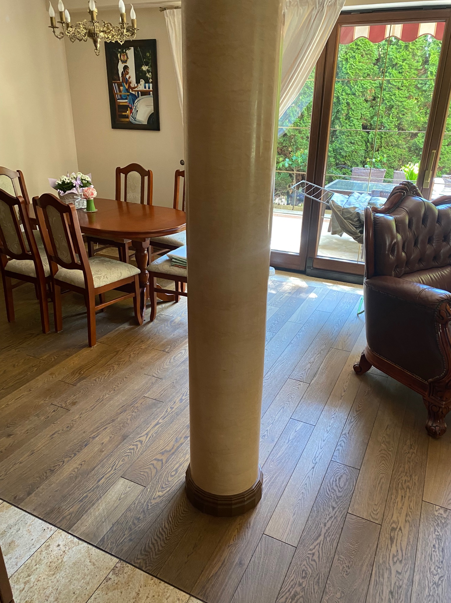 Jasne drewniane panele podłogowe w jadalni z kolumną, brązowy skórzany fotel i widok na zieleń przez okno. Ciepłe oświetlenie wnętrza.