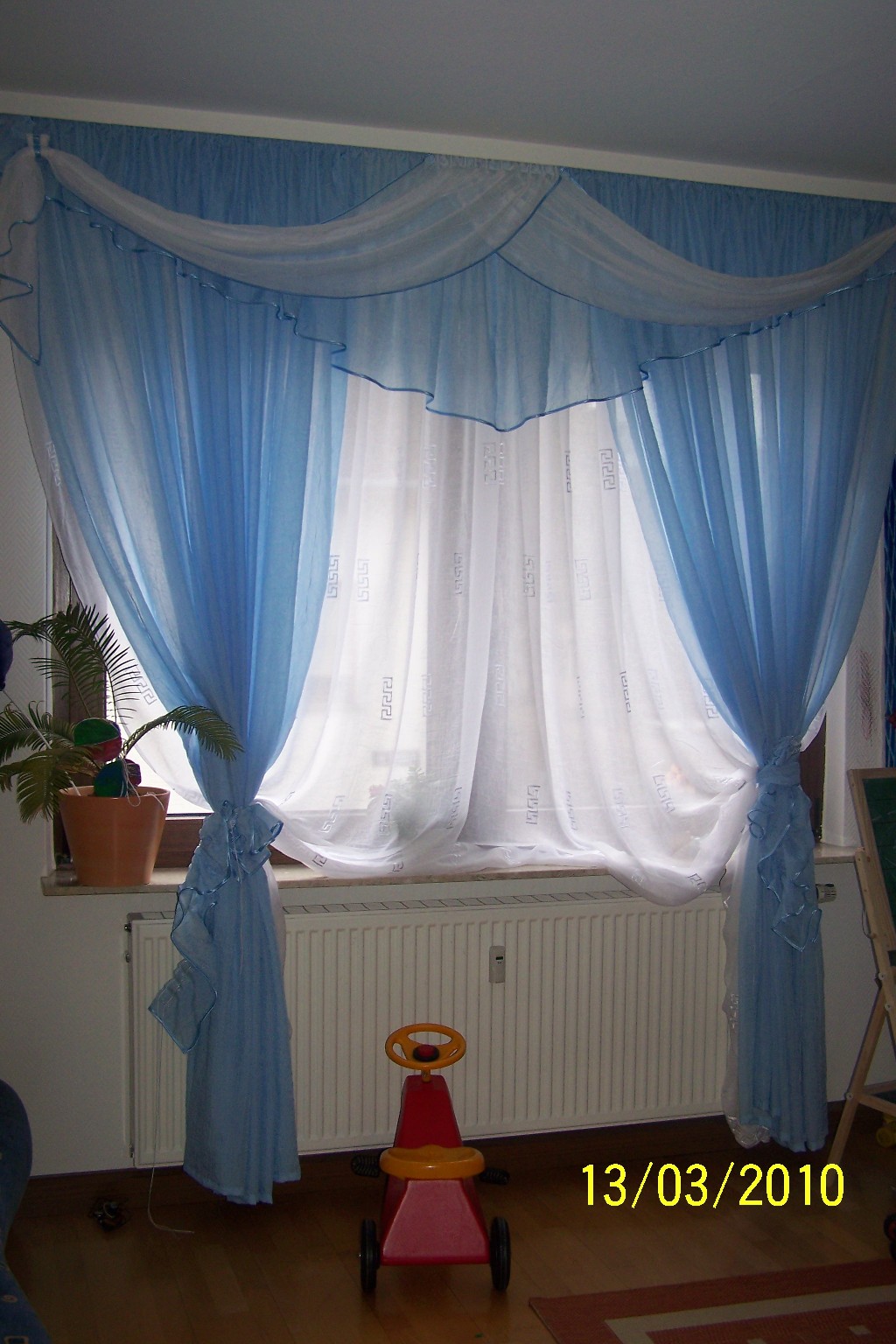 Okno udekorowane błękitno-białymi, zwiewnymi zasłonami z falbanami, upiętymi po bokach, z rośliną doniczkową na parapecie i dziecięcym jeździkiem pod oknem.