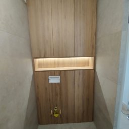 Meble Localo - Wysoka zabudowa z drewnianymi frontami w toalecie, z podświetlaną wnęką, ukrywająca instalację sanitarną, na tle beżowych płytek.