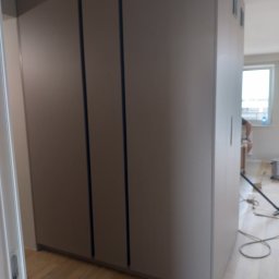 Meble Localo - Duża, szara szafa wnękowa z przesuwnymi drzwiami, widoczna w trakcie montażu, z narzędziami i pracownikiem w tle.