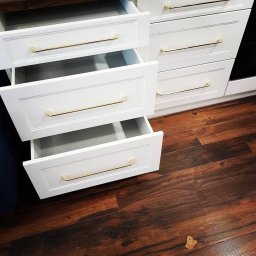 Meble Localo - Białe szuflady z frezowanymi frontami i złotymi uchwytami, dwie otwarte, na tle ciemnej, drewnianej podłogi.