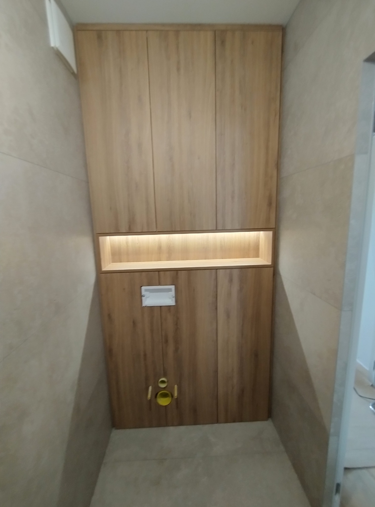 Wysoka zabudowa z drewnianymi frontami w toalecie, z podświetlaną wnęką, ukrywająca instalację sanitarną, na tle beżowych płytek.