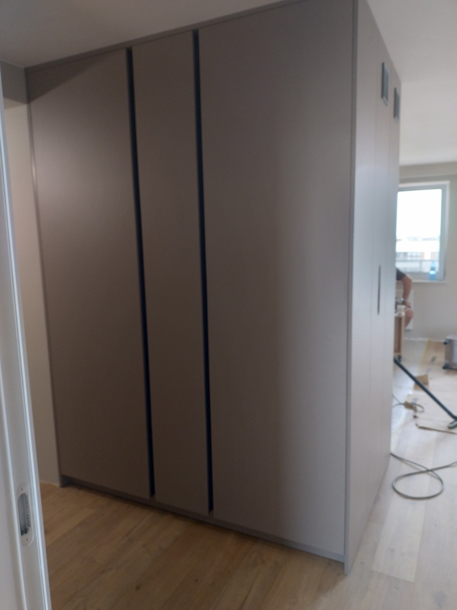 Duża, szara szafa wnękowa z przesuwnymi drzwiami, widoczna w trakcie montażu, z narzędziami i pracownikiem w tle.