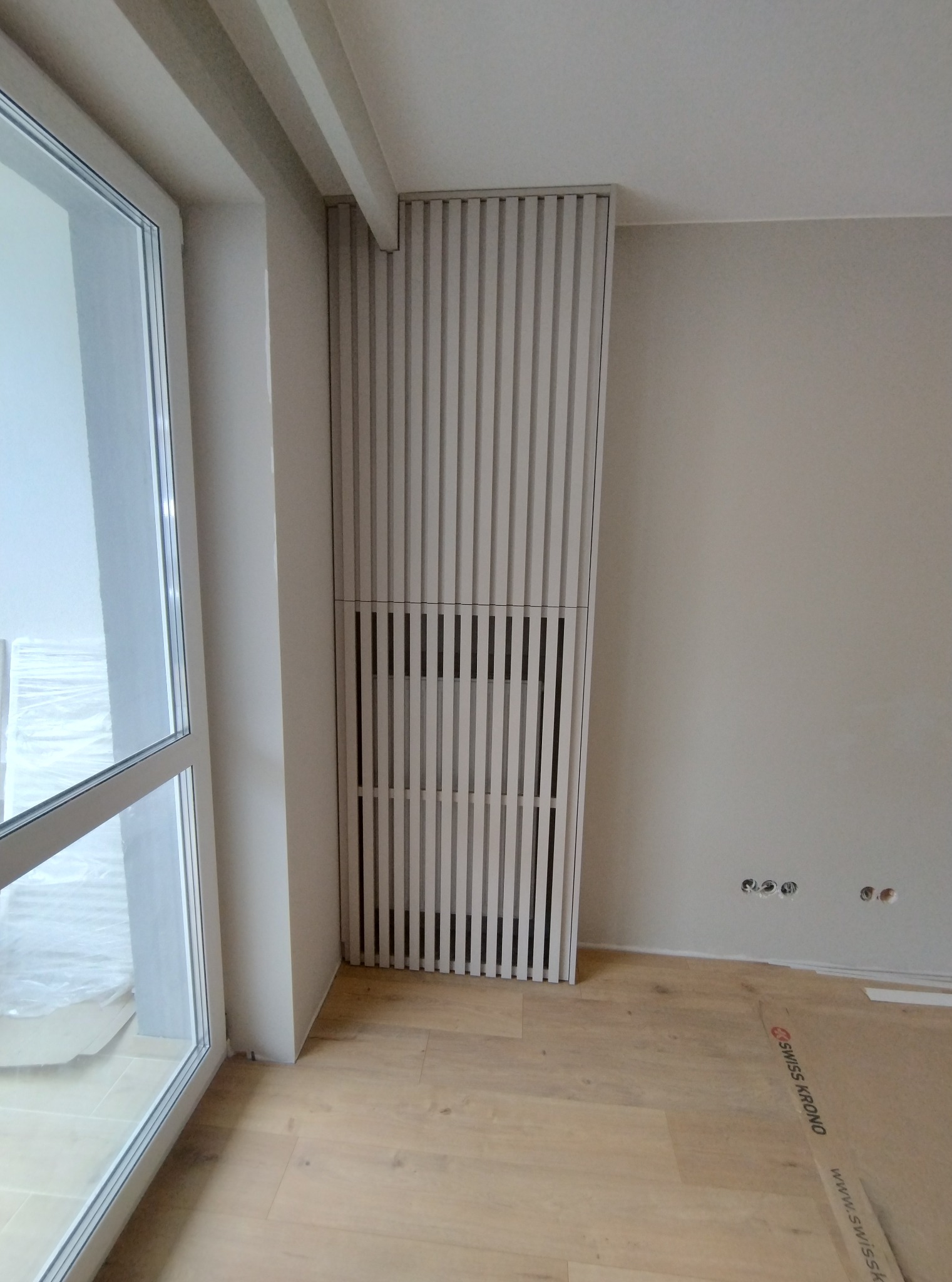Biała, pionowa szafa wnękowa z lamelami, częściowo zakrywająca grzejnik, widoczna na tle jasnej ściany i podłogi z paneli, obok okna.