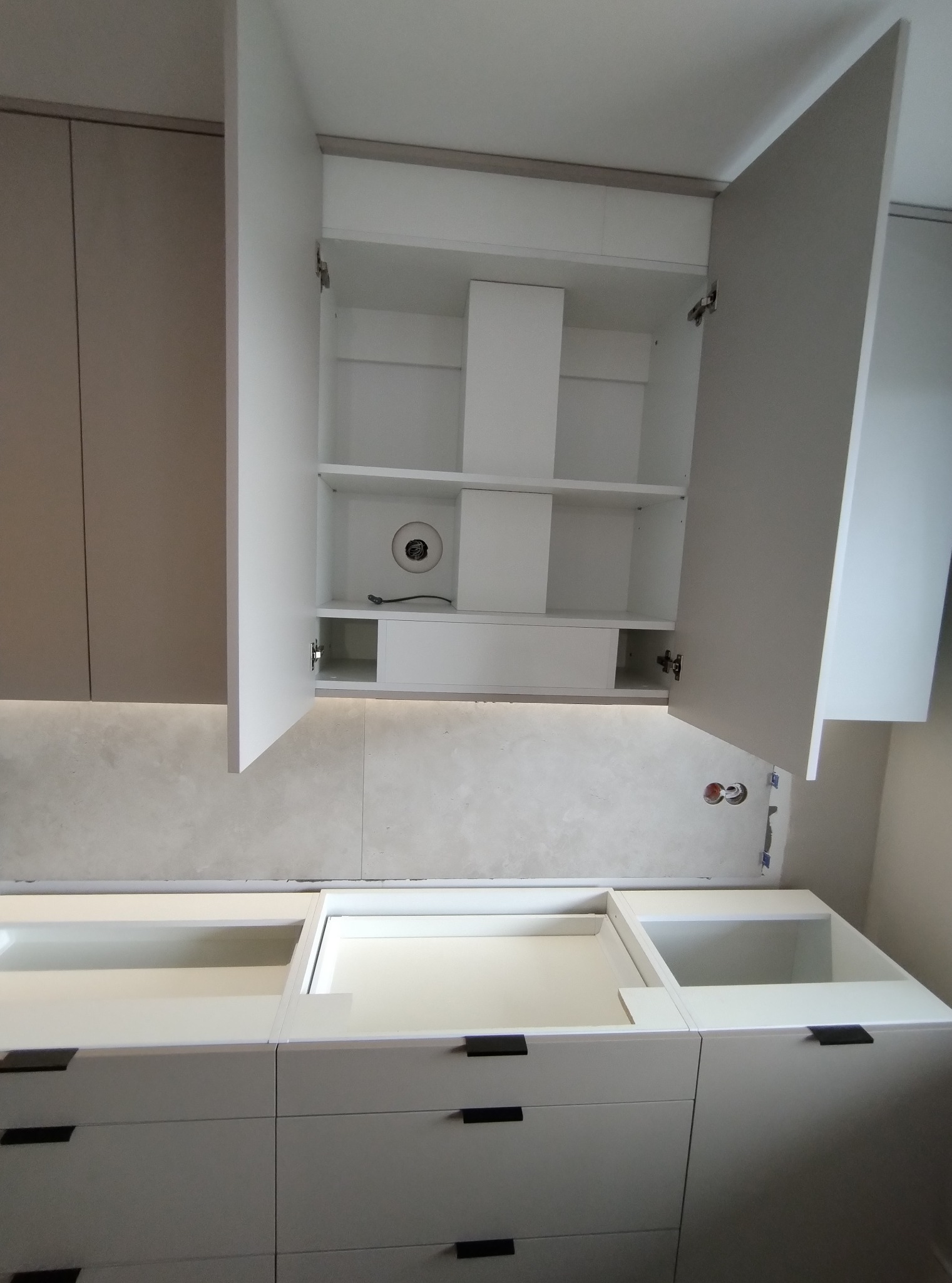 Montaż białych szafek kuchennych z otwartymi górnymi szafkami, widoczne okablowanie i otwór na rurę wentylacyjną w jednej z szafek wiszących.